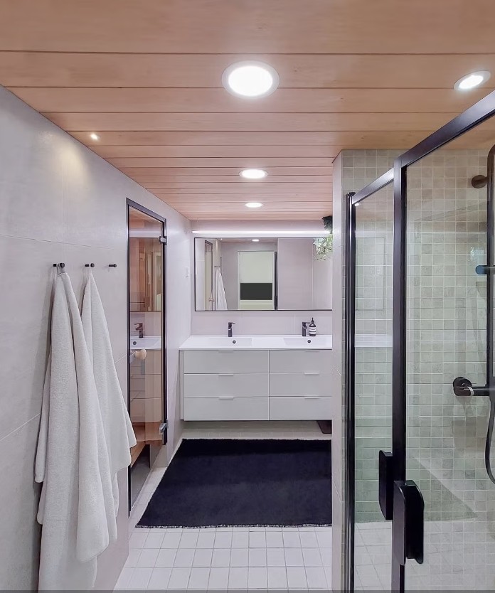 A modern bathroom with a double sink, white cabinetry, a glass shower area, and a warm wooden paneled ceiling. Soft towels hang from hooks on the left wall. The stylish ensemble is completed with energy-efficient LED lighting from LedStore, which enhances the modern look of the bathroom and creates ambiance throughout the space. - LedStore Combinations