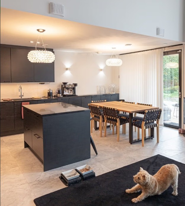A modern kitchen and dining area with black cabinets, a wooden dining table, and comfortable LED lighting from LedStore.fi create a cozy atmosphere. A cat stretching on the black rug completes the homely look. - LedStore A modern kitchen and dining area with black cabinets, a wooden dining table, and comfortable LED lighting from LedStore.fi create a cozy atmosphere. A cat stretching on the black rug completes the homely look.