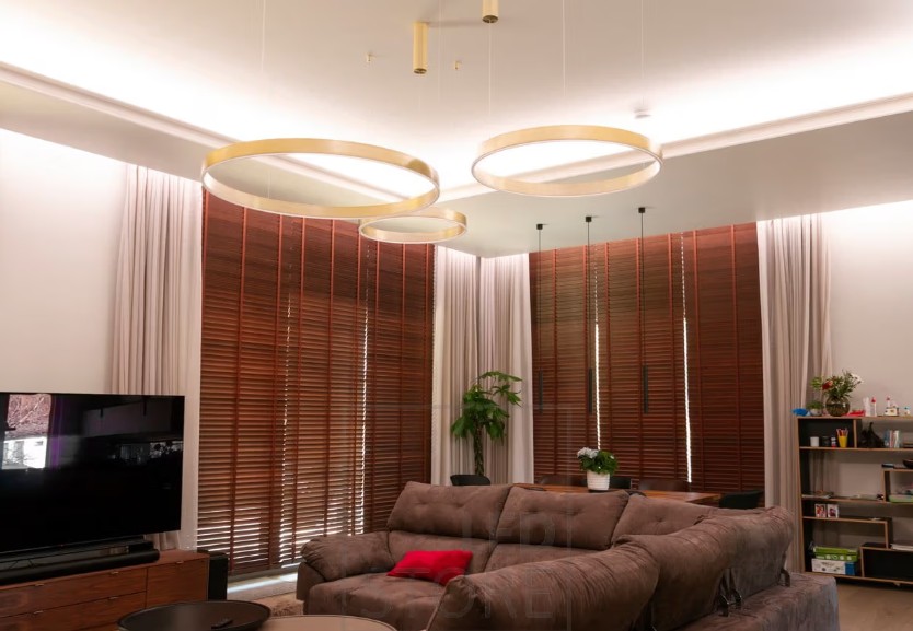 Moderni olohuone, jossa ruskeat sälekaihtimet, ruskea kulmasohva, tyylikkäät kultaiset led-rengasvalaisimet sekä televisio vasemmalla. - LedStore In the living room, the indirect LED strip lighting is installed along the ceiling and in front of the curtains, casting light downward. Stylish decorative lights are added for extra flair.