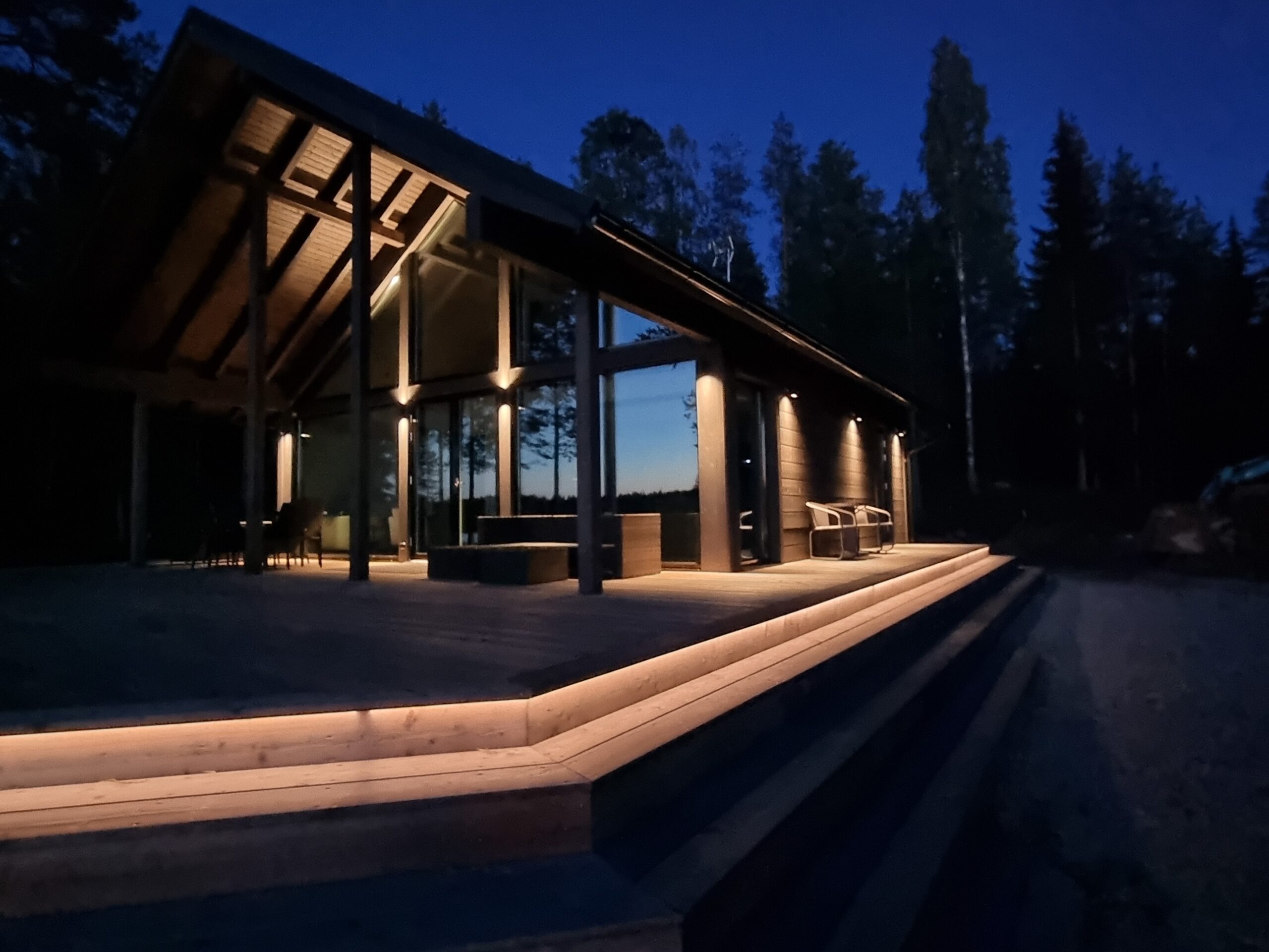 A modern log cabin with large windows and outdoor stairs is bathed in warm ambient light, thanks to high-quality outdoor lighting and LED strip lights from LedStore.fi. Stylish LED fixtures highlight the cabin’s architecture and surrounding nature, creating an inviting atmosphere at dusk. - LedStore LED strip lights can be installed over long distances as a continuous line. In this example, the steps surrounding the terrace are illuminated so that an LED strip is placed under the first step. The contrast created by the light clearly indicates where the first step begins.