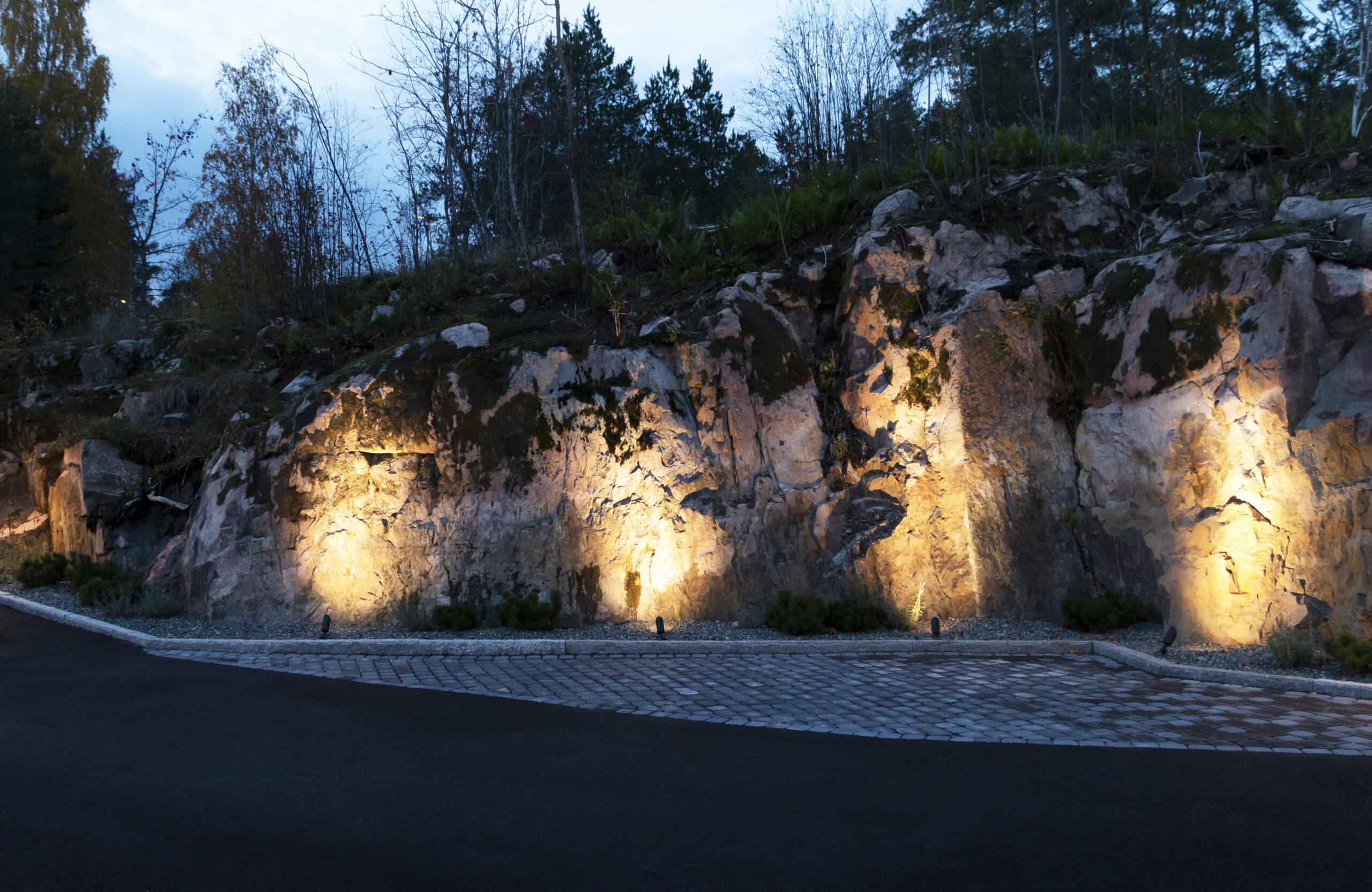 At the edge of the rocky yard, LED spike lights highlight the rock wall with upward-facing light from below.