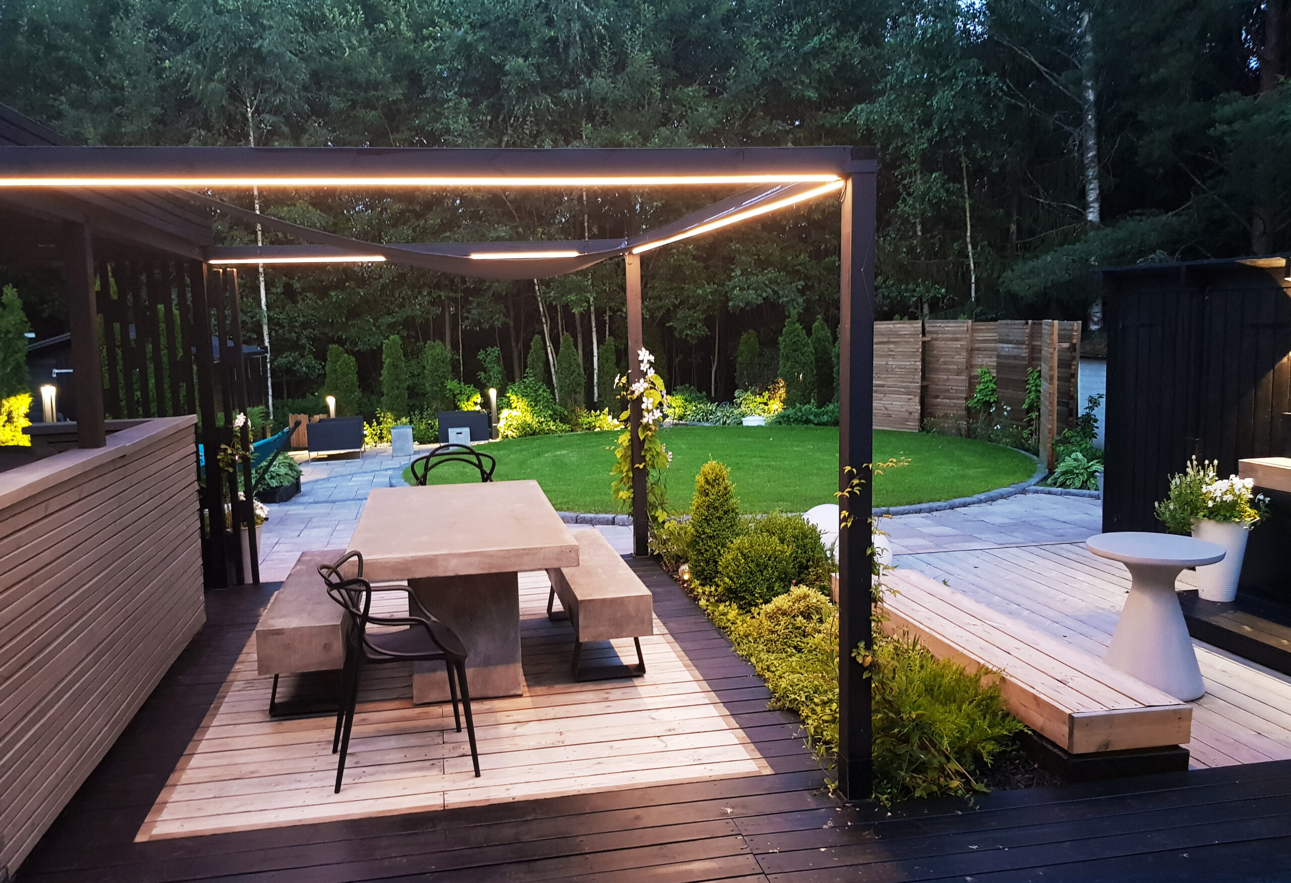 A modern backyard terrace with a pergola, dining table, and lush garden, where the well-kept lawn takes on a stunning look in the fading evening light as LedStore.fi’s outdoor lighting and stylish LED strip lights create a cozy, atmospheric glow. - LedStore LED strips in aluminum profiles installed along three edges of the canopy frame above the dining area in the outdoor kitchen.