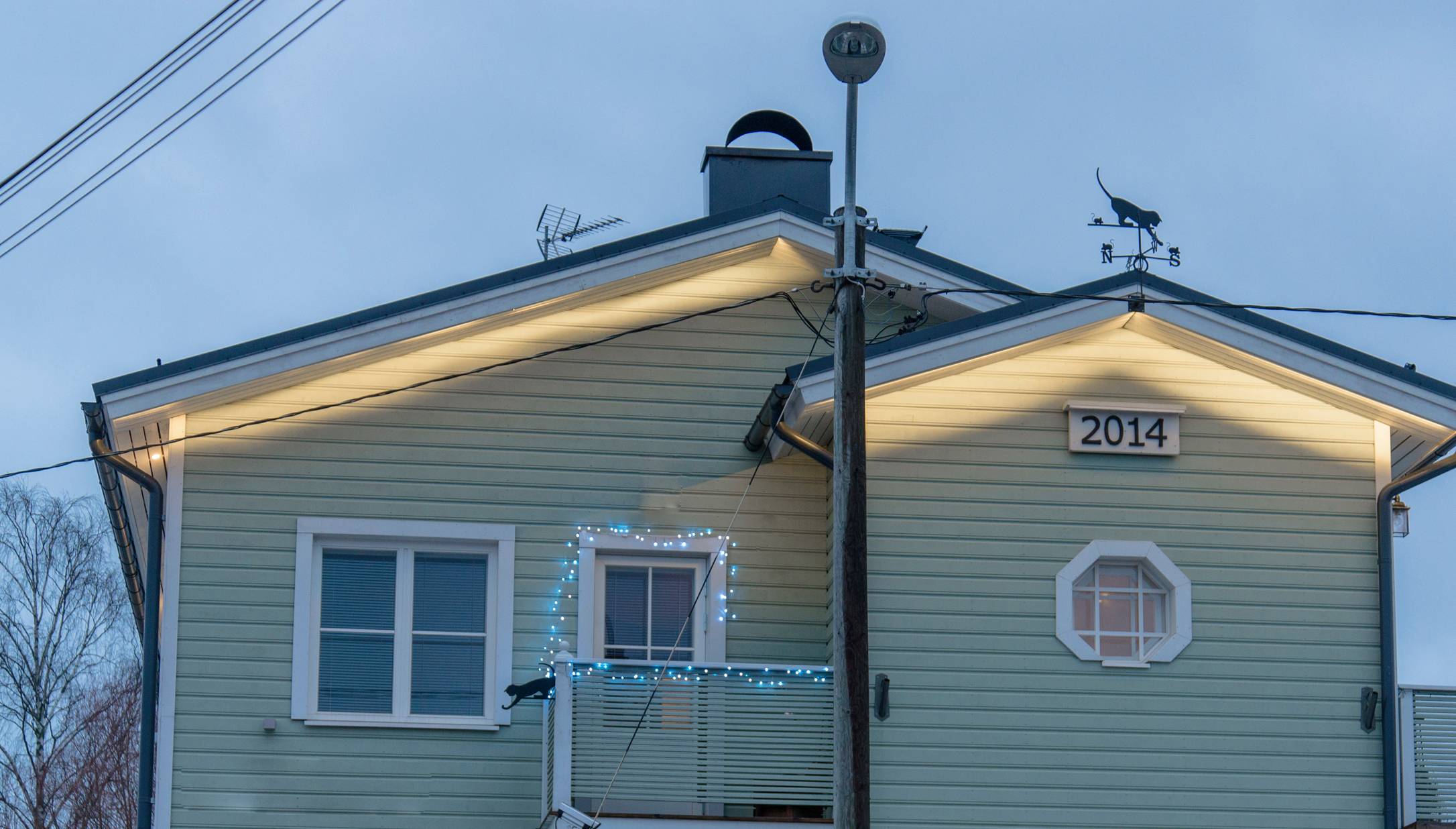 A light green wooden house with the year "2014" on the facade. For outdoor lighting, atmospheric strips and weatherproof light sets from LedStore.fi are used, with LED lighting on the balcony. A cat-shaped weather vane decorates the roof. - LedStore LED strips used in outdoor lighting at the end of a pitched-roof house in two different configurations.