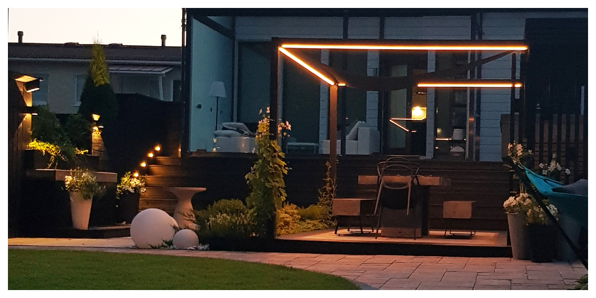A modern backyard patio with a pergola and stunning LED mood lighting. A wooden staircase and atmospheric potted plants complete the look. This setup, located in the yard of a modern home, offers the perfect setting for evening moments, all enhanced by LedStore.fi's high-quality LED lighting. - LedStore Backyard patio with a pergola lit with LED strip. A wooden staircase illuminated from the side.