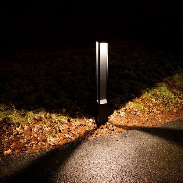 Outdoor LED light post illuminating a path with warm glow, highlighting fallen leaves and pavement texture; darkness surrounds the lit area.